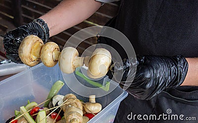 Ð¡hef cooks mushrooms. Man in cooking uniform and gloves prepare champignon skewers for grilling. Outside picnic party kitchen Stock Photo